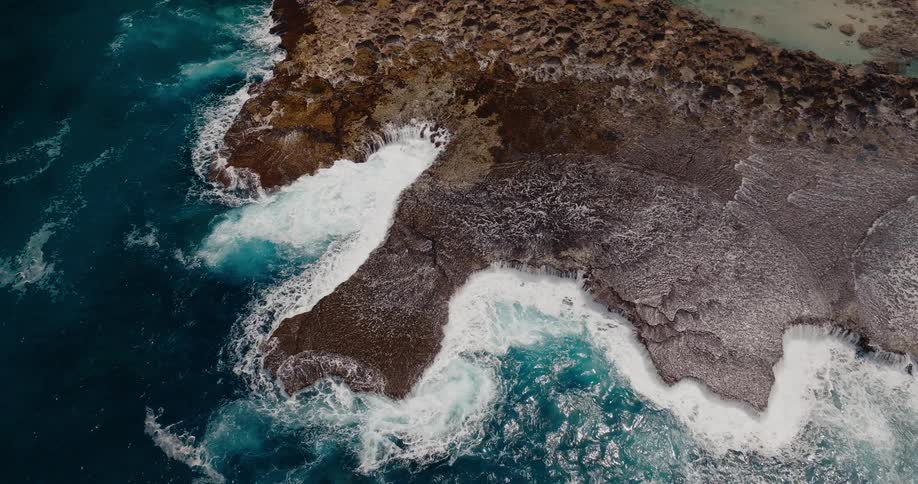 High aerial view showing powerful turquoise waves crashing violently onto a dark brown rugged rock shelf creating dramatic arcs of white sea foam