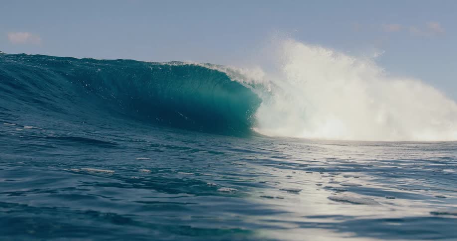 A massive deep blue ocean wave curls and crashes into white spray under a bright blue sky filmed from a low perspective on the surface of the water