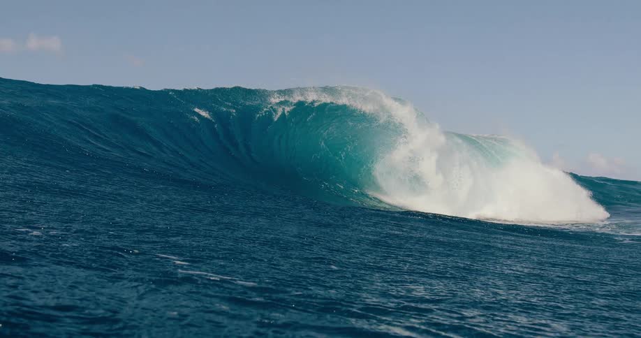 Low angle view shows a massive dark blue ocean wave building up and breaking violently creating a white foamy barrel under a clear sky