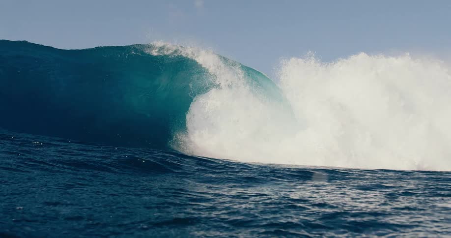 Powerful deep blue wave curls and crashes in the open sea under a blue sky with clouds as the view becomes submerged in the water
