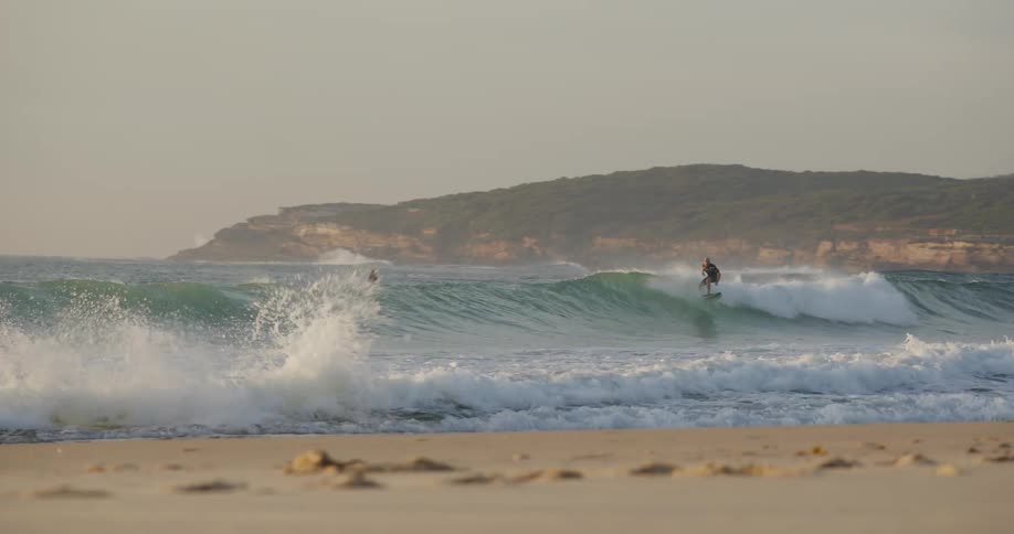 A man wearing a black wetsuit surfs a large green wave toward the shore on a beach with a blurred foreground and a coastal cliff in the background