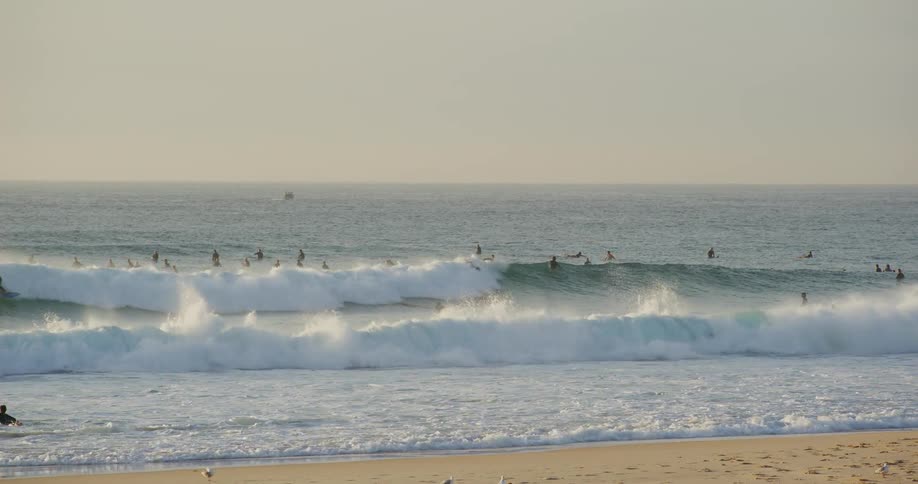 Dozens of surfers wait on their boards in the ocean beyond the foam of breaking waves near a sandy beach under a bright hazy sky