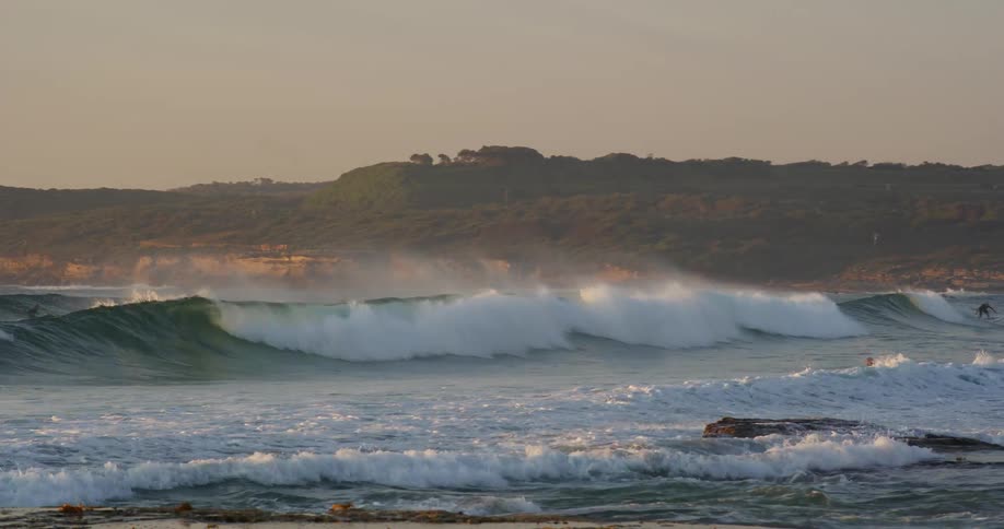 Surfers ride rolling waves near a rocky shore bathed in the warm light of a golden sunset with a coastal hill in the background