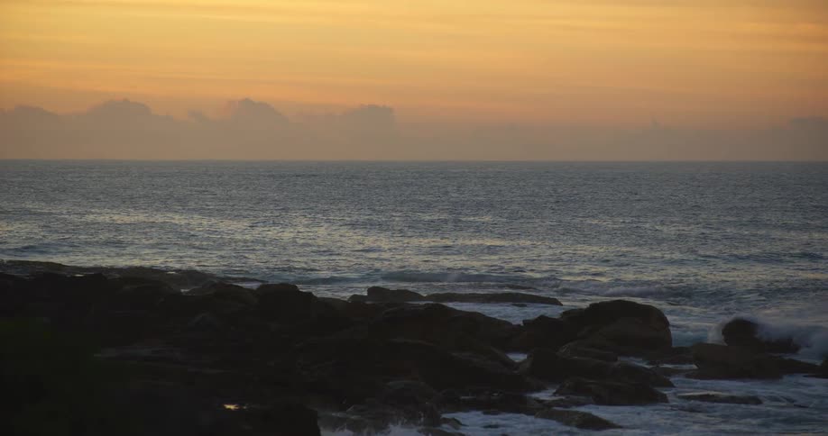 A wide view captures the dark silhouette of a rocky shoreline with crashing waves under a bright orange and gray sunset sky