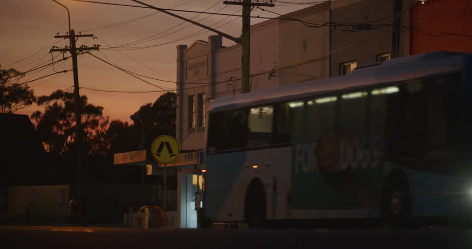 A city bus with bright headlights passes by low rise commercial buildings in the late evening with an orange sky and utility poles