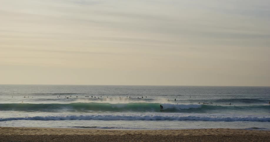 Silhouettes of surfers ride and wait for waves on the ocean water near a sandy beach as gentle waves roll toward shore under a pale sky