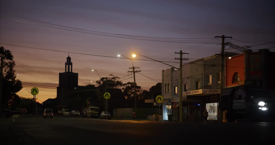 Urban street at dusk with moving cars and storefronts lit by streetlights