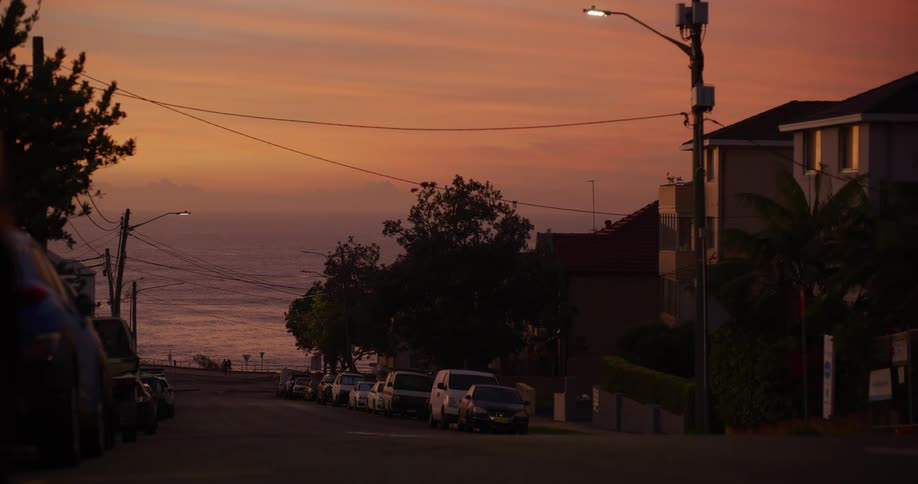 Cars parked on a neighborhood street as light traffic moves and people walk along the coast and a man crosses the street in front of a bus