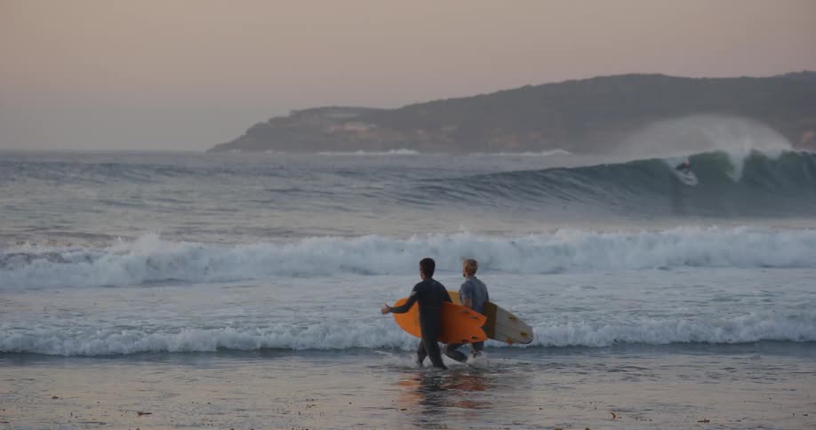 Two men wearing dark wetsuits walk into the rolling ocean waves holding surfboards on a beach at dusk with a large hill in the distance