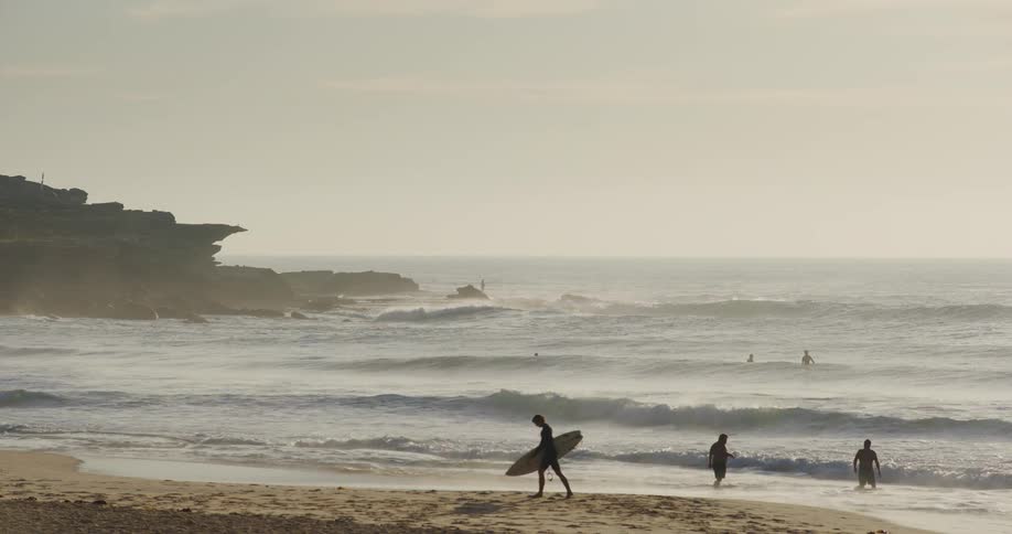 A person walking along the beach carrying a surfboard while nearby tourists enjoy swimming in the ocean