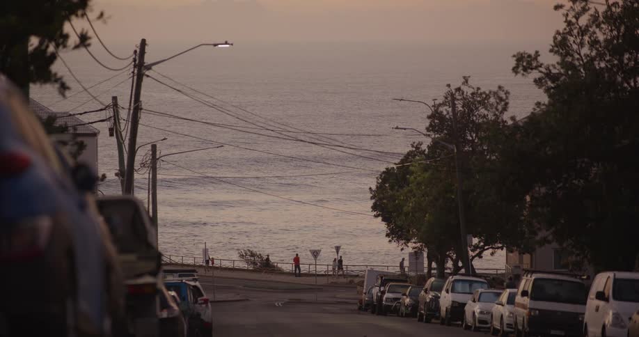 Cars parked on a neighborhood street as light traffic moves and people walk along the coast