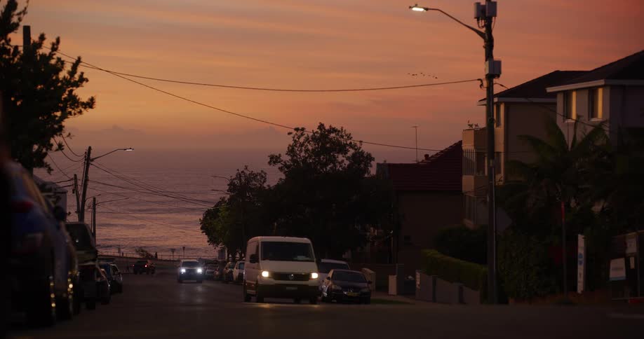 Cars parked on a neighborhood street as light traffic moves and people walk along the coast