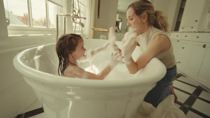 Mother and daughter play as the child sits in a bubble bath during the daytime