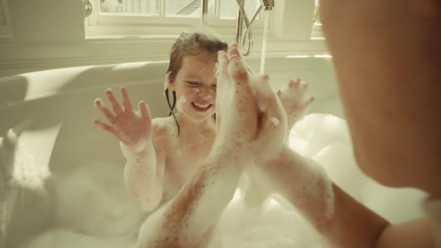 A playful scene where an adult leans over a bathtub laughing while helping a young child covered in wet hair and foam play with the large bubbles in a sunlit bathroom