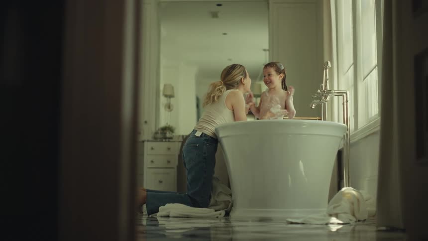 Woman kneeling beside a bathtub interacting with a child in a welllit bathroom