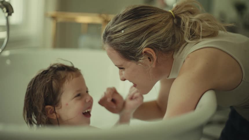 Mother playfully bathing her daughter in a bathtub indoors during daytime