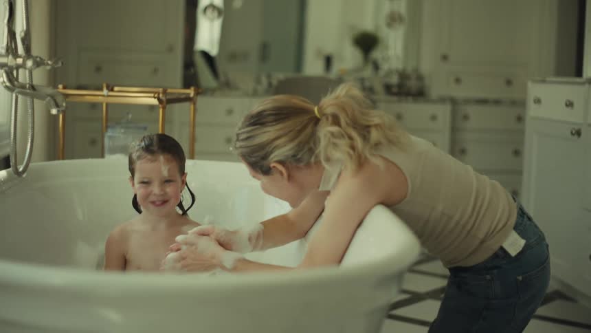 Young adult woman playing with foam while a small girl inside a bathtub laughs indoors during day time