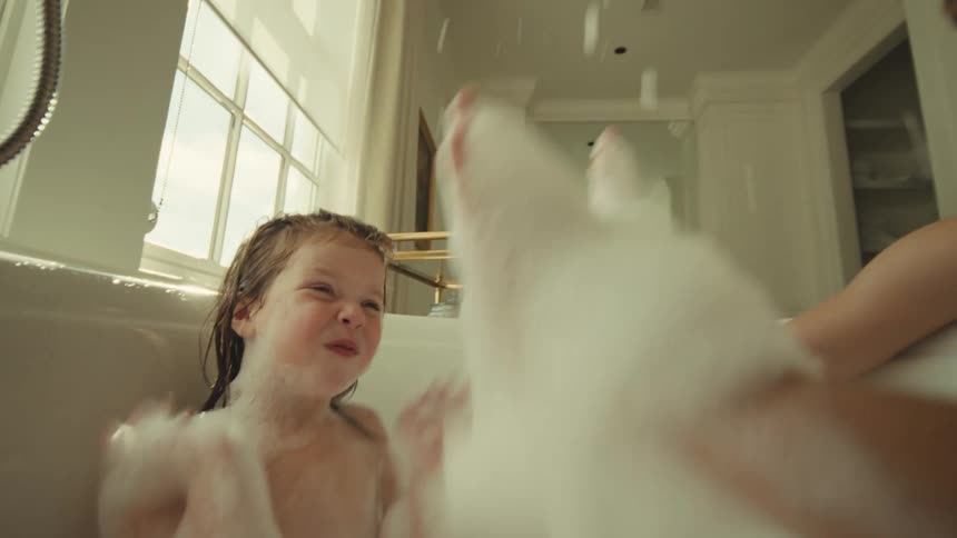 A playful scene where an adult and a young child are taking a bubble bath in a bright luxurious bathroom laughing as the child interacts with the large amount of soap foam