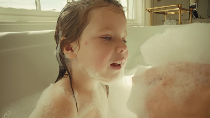 Girl on a bathtub blowing at soap foam on the hands of another person while smiling indoors during day time