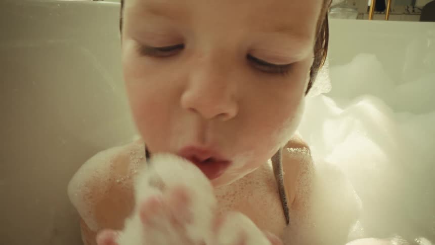 Child in a bathtub holding foam while a hand adds more foam to the bath
