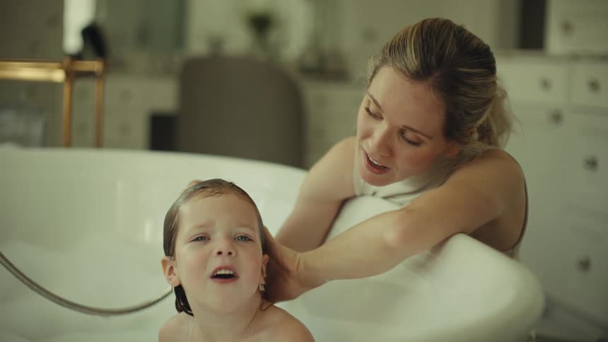 Young adult woman washing the hair of a small girl in a bathtub while they talk and smile indoors during day time