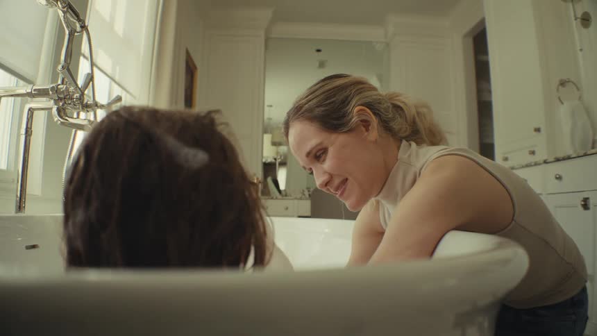 Young adult woman washing and talking to a girl on a bathtub while she plays around with the foam and both smile indoors during day time