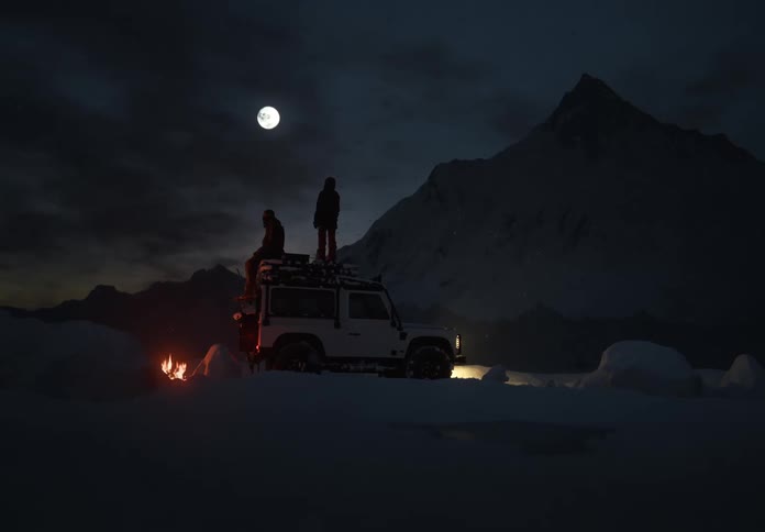 Two people on top of an expedition 4x4 at night with a campfire and full moon in snowy mountains, winter adventure scene.