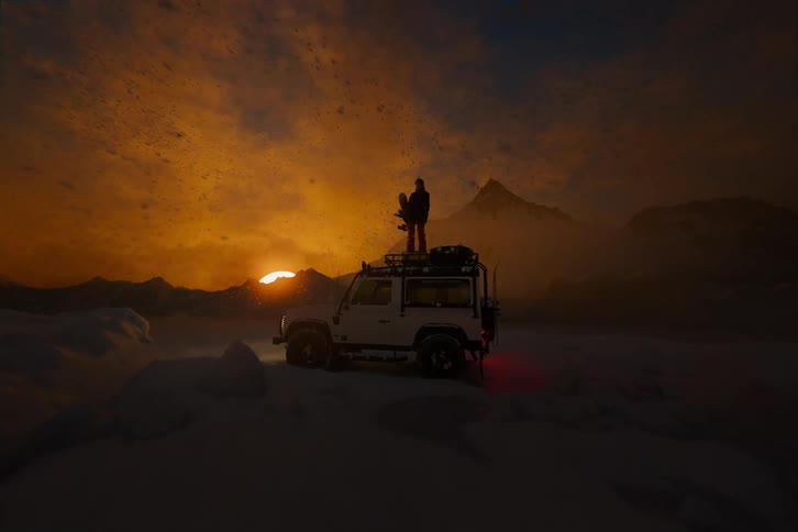 Woman with a snowboard standing on an off-road SUV during a golden hour winter snowstorm, blowing snow in mountains.