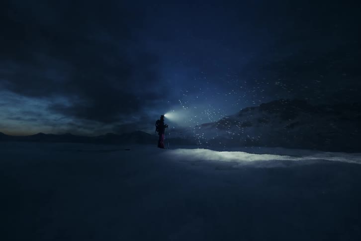 Hiker wears a headlamp as he stands in the falling snow holding trekking poles