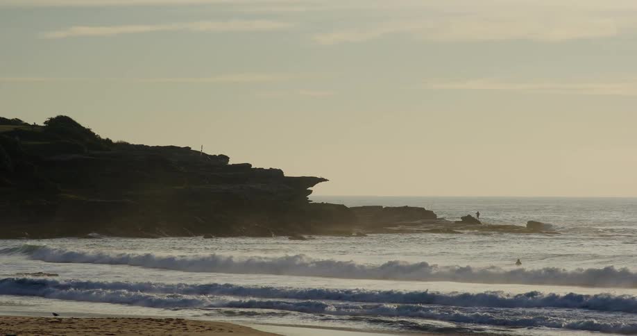 Person stands on the rocks near the water while surfers enjoy the waves on a cloudy day