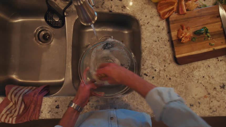 This video shows a person washing a clear glass mixing bowl in a stainless steel kitchen sinkusing an overhead view the person uses a lightcolored scrub brush and running water to clean the bowl before rinsing it clean