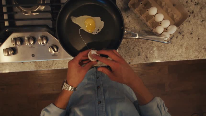 A person wearing a denim shirt cracks an egg into a pan on a stove top above a granite kitchen counter