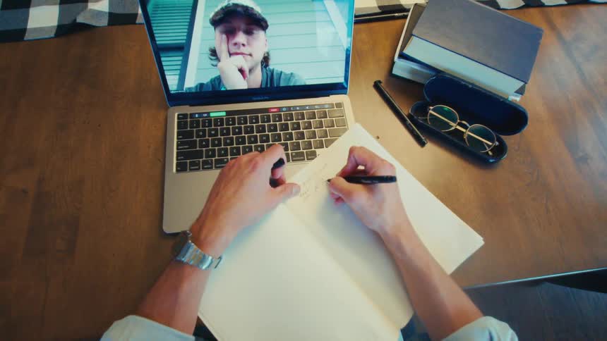 Person writing in a notebook while participating in a video call on a laptop