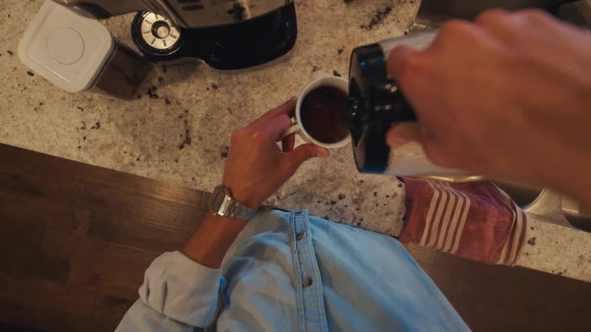 A person pours coffee from a coffee maker into a mug and enjoys it while in the kitchen