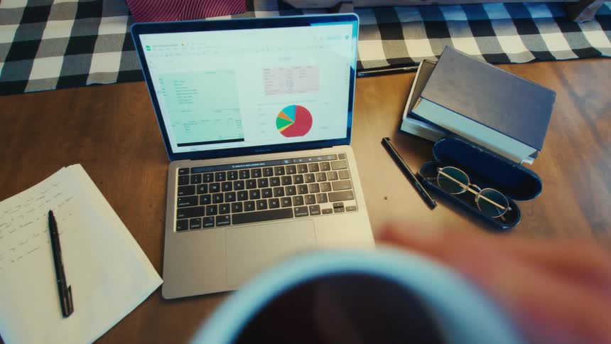 A person wearing a silver watch works diligently on a laptop displaying data and a pie chart on a wooden desk occasionally pausing to take a sip from a white coffee mug surrounded by books a notepad and glasses in a case