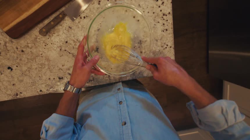 A man wearing a denim shirt and a watch is seen from a first person perspective whisking eggs in a glass bowl on a marble kitchen counter
