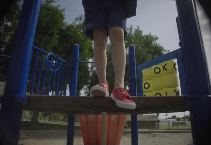 A child wearing red sneakers and blue shorts climbs onto the platform of a blue and orange playground structure with a yellow tictactoe game board visible nearby outdoors