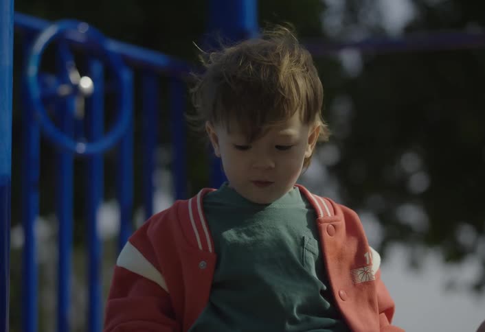 A little boy is sitting in a playground swinging his legs and wearing red sneakers