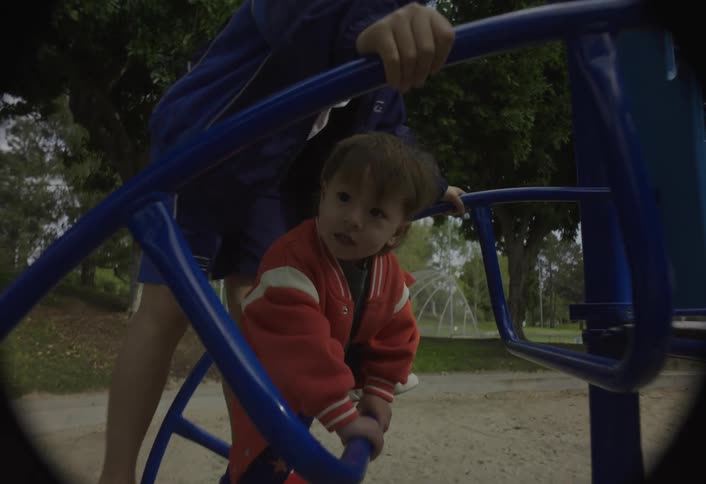 This video shows a short scene at a park playgrounda young child in a red and white jacket is climbing a blue metal structure assisted by an older person in a dark blue jacket and a baseball cap