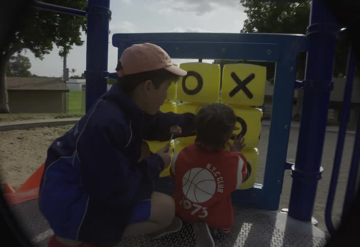 The two children are now interacting with a large yellow rotating tictactoe game board on the playground structurethe older child is guiding the younger childs hand to turn one of the cubes revealing a black o