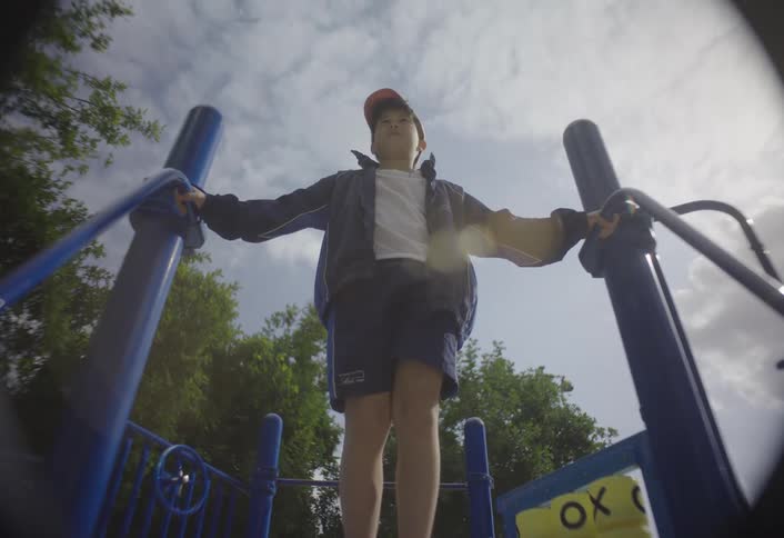 The video filmed from a lowangle perspective shows a young boy standing on a blue wooden or metal play structure at a playground he is wearing a dark jacket dark shorts a white shirt a red baseball cap and red and white sneakers and he appears to