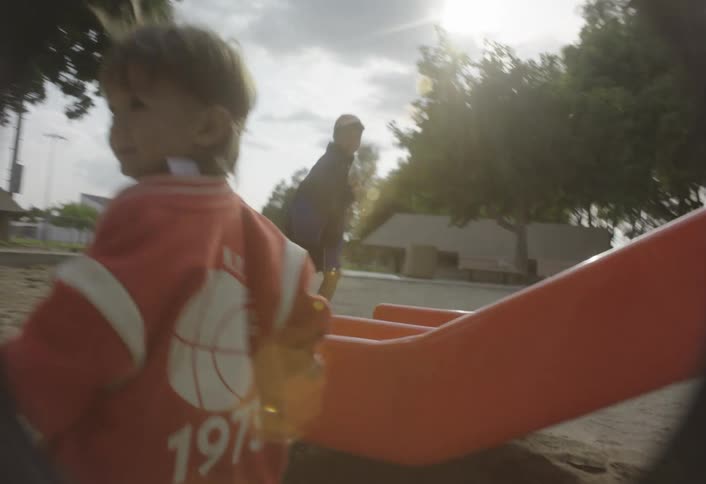 A young child wearing an orange jacket and blue pants runs across the sandy ground of a playground passing by a bright orange slide as seen from a low wideangle perspective