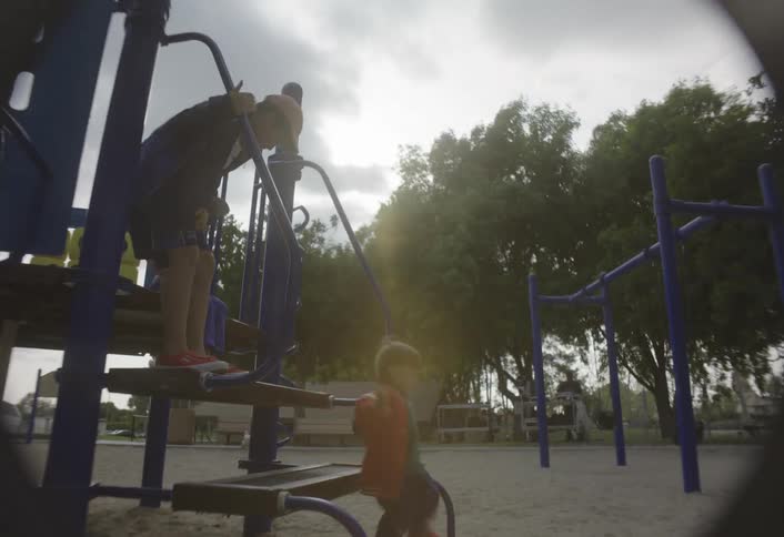 Through a distinctive fisheye lens a toddler in a red jacket climbs down from a blue playground structure and toddles across the sand while two older children watch from the platform above