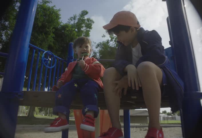 Boy smiles at the younger boy as the younger boy eats a lollipop while they sit on playground equipment