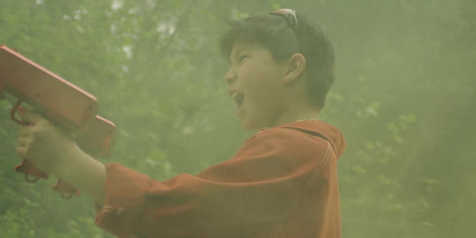 A young boy in an orange jacket and gold chains screams excitedly while holding two red toy money blasters that rapidly spray fake dollar bills around him in a hazy green outdoor setting