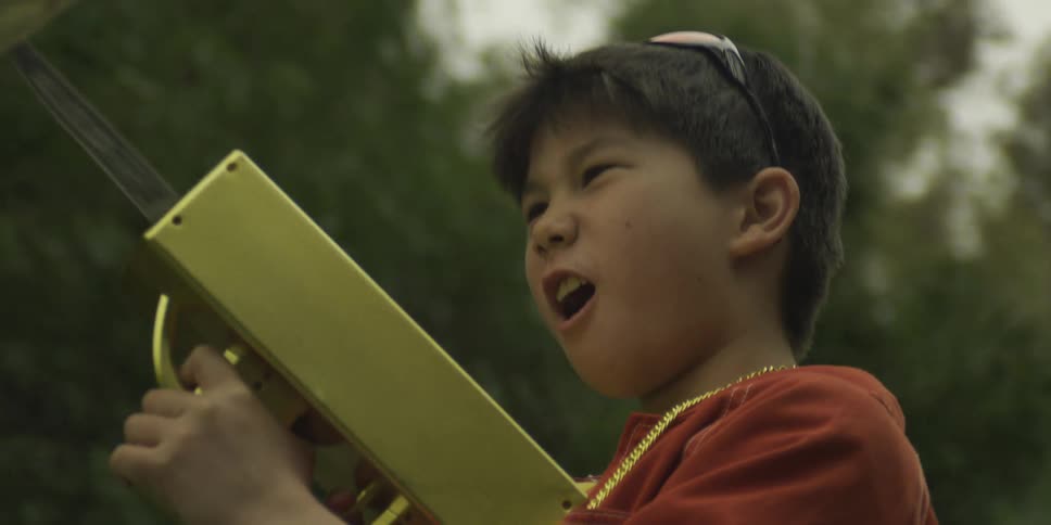 With a joyful shout a young boy in a red jacket aims a golden toy gun at the sky spraying a cascade of fake money into the air around him
