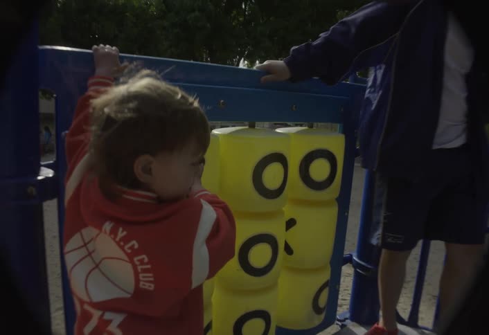 Two children one in a red hoodie and one in a cap are on a playground structure with a yellow tictactoe game the older child talks while the younger child climbs down the games rotating blocks