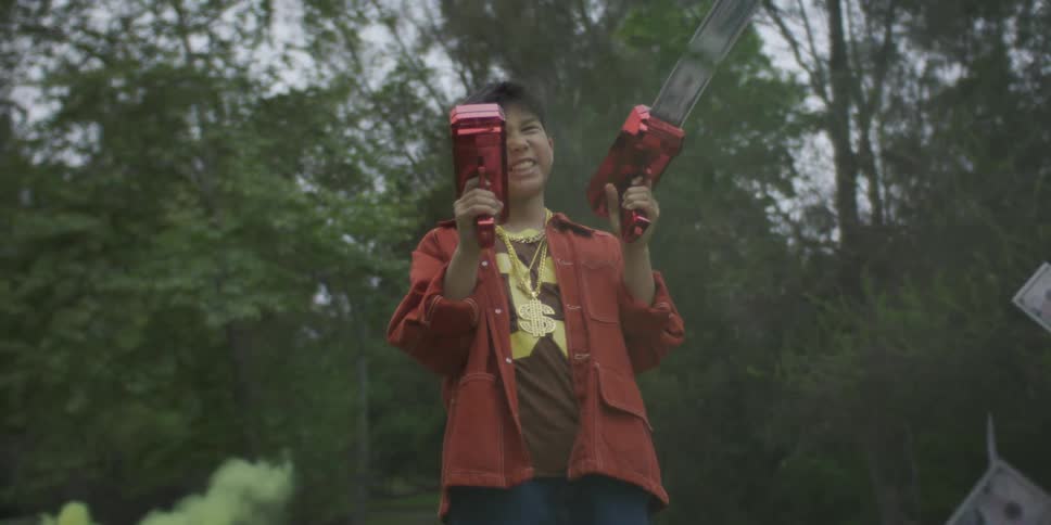 A boy wearing a gold chain and red jacket stands in a park swirling with green smoke enthusiastically firing two red toy guns that shower the air with fake cash