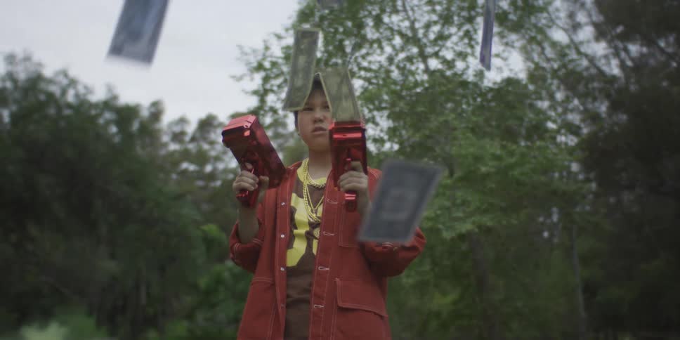 Young boy standing outside holding two red toy guns shooting out hundred dollar bills