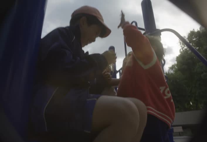 Three asian children an older one wearing a cap a younger one in a red jacket and a young girl are playing and interacting closely while sitting and standing on a blue metal play structure at a park